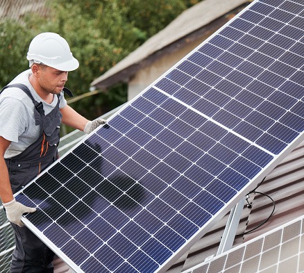 Man technician carrying photovoltaic solar moduls on roof of house. Electrician in helmet installing solar panel system outdoors. Concept of alternative and renewable energy.